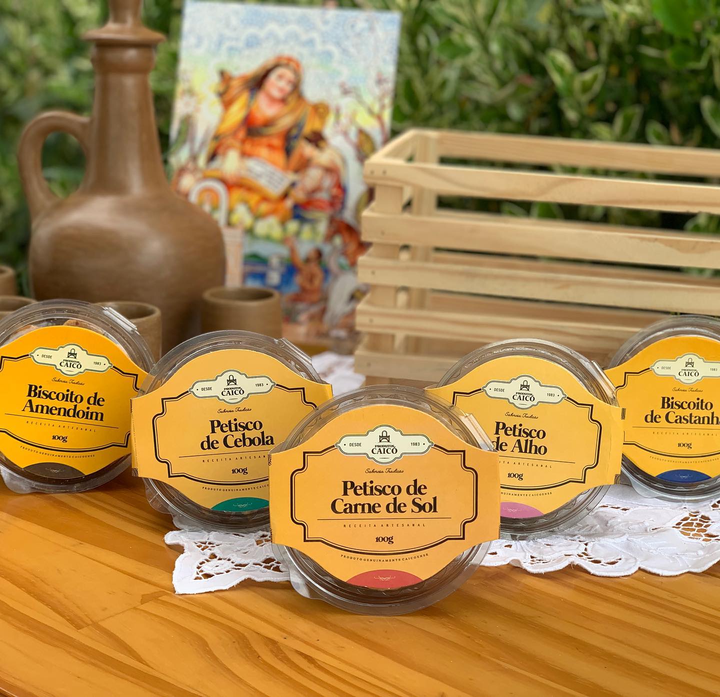 Mesa farta com biscoitos e petiscos artesanais Produtos Caicó, cercada por um jarro de barro e imagens de devoção, a mesa de uma casa caiçoense.