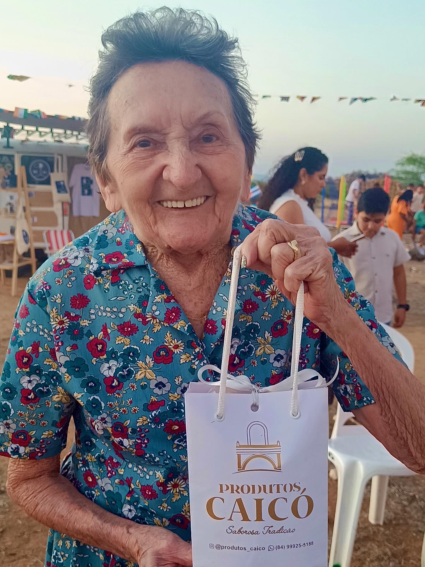 Tânia Maria sorridente segurando uma sacola da marca Produtos Caicó em uma festa junina no Seridó